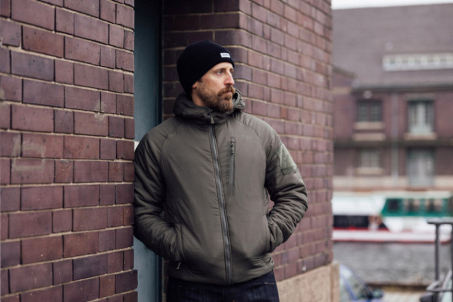 Mann mit Helikon-Tex Winterjacke und Mütze lehnt an Backsteinwand in der Stadt