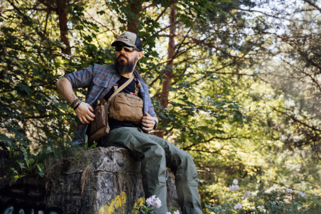 Mann mit Helikon-Tex Chest Rig und Outdoor-Bekleidung sitzt auf Felsen im Wald