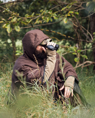 Jäger in braunem Poncho mit Fernglas bei der Beobachtung – präzise Ausrüstung für verantwortungsbewusstes Weidwerk.