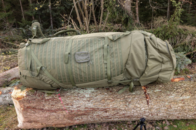 Übergroßer olivfarbener TT Mega Mil Ops Pack 600 liegt auf einem Baumstamm im Wald, mit Tragegriffen und Befestigungsschlaufen sichtbar.