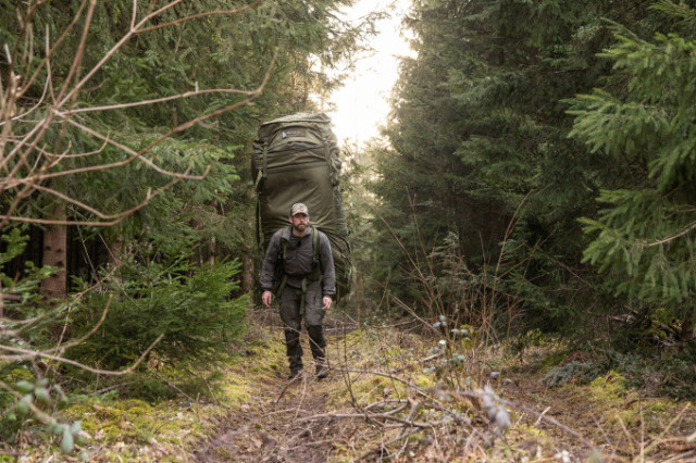 Person läuft mit riesigem olivfarbenen Rucksack durch Waldgelände.
