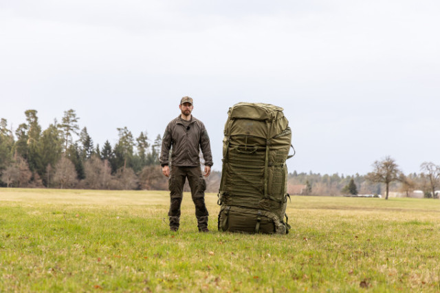 Man steht neben einem extrem großen olivfarbenen Rucksack auf freiem Feld.