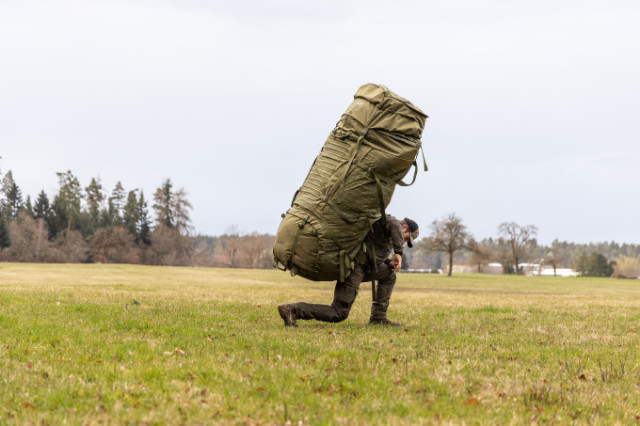 Man steht neben einem extrem großen olivfarbenen Rucksack auf freiem Feld.