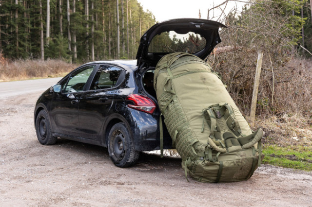 Man steht neben einem extrem großen olivfarbenen Rucksack auf freiem Feld.